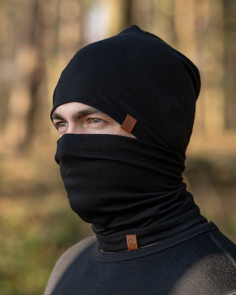The Menique Men's Merino Beanie and Gaiter Set in Black color. The man is wearing the accessory set in a forest, in fall time.
