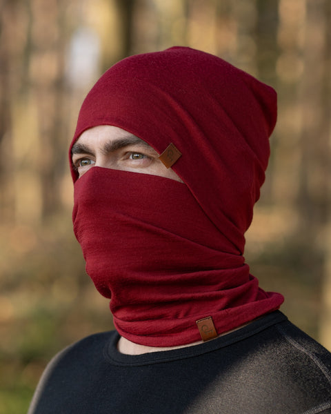 A man, standing outside in a forest during fall season, wearing the Menique Men's Merino Beanie and neck Gaiter Set in the color Royal Cherry.