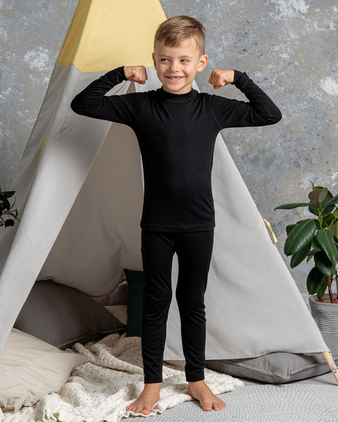 a boy wearing the menique Kids' Long Sleeve Set 160 Merino in Black color. He is standing with his arms raised ant bent, acting strong.